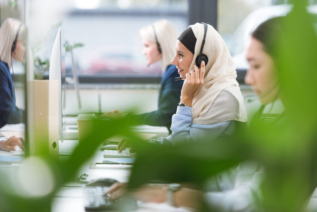 selective focus of smiling arabic call center operator in hijab in headset working on computer at